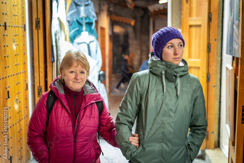 Senior mother and adult daughter walking through a colorful shopping street at night in Tangier, Morocco, smiling at the camera and enjoying their winter holiday together.
