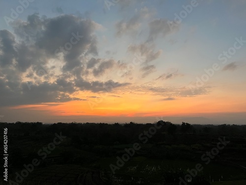 Fotografie Vibrant orange sunset with dramatic clouds over rural fields and distant hills, scenic evening sky in countryside