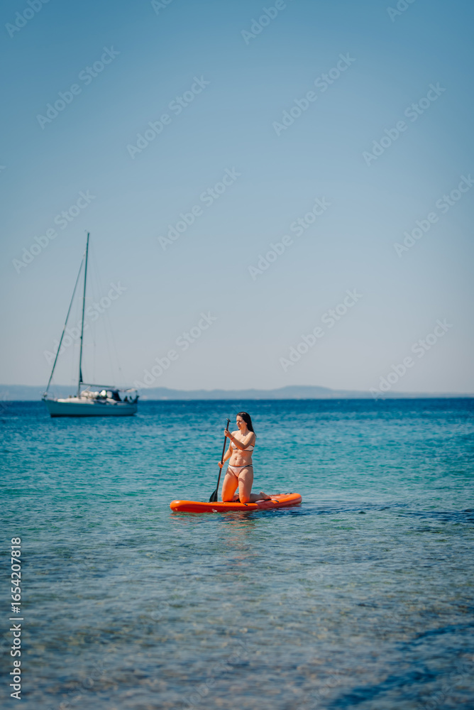 Naklejka premium Woman kneeling on sup board paddling in the sea with sailboat in background