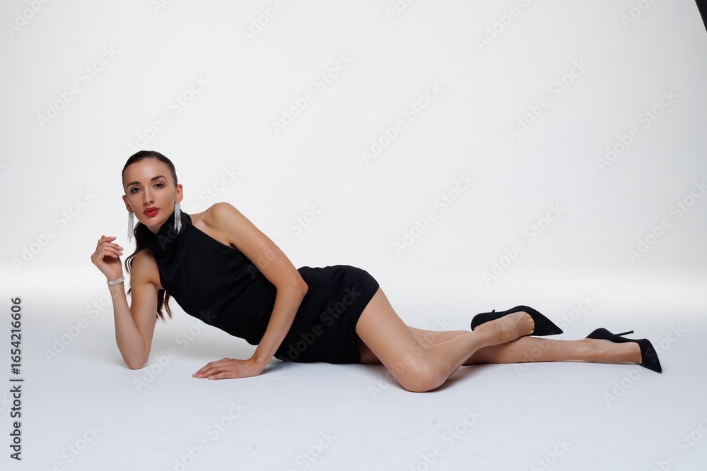 Obraz premium Beautiful young woman in a black elegant dress posing in studio on white background. Fashion model with long hair, red lips, and stylish earrings. Glamour portrait, studio photoshoot, beauty 