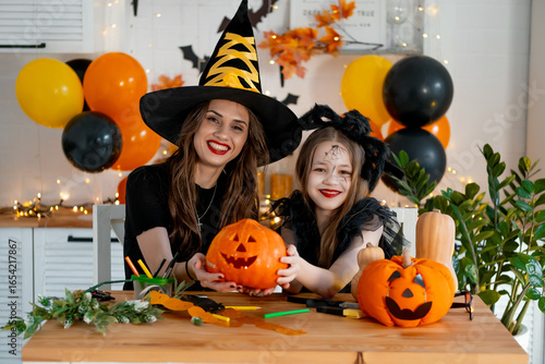 A happy family is getting ready for Halloween. Mother and child daughter in carnival costumes at home in the kitchen have fun painting and carving pumpkins and congratulating on the holiday