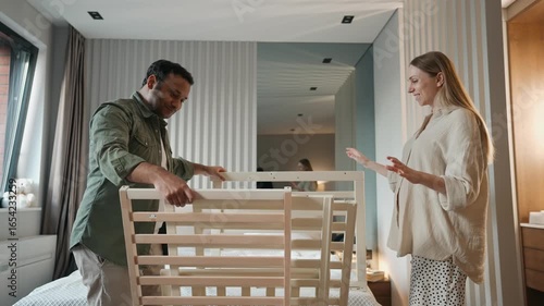 A pregnant woman approaches a man who is holding parts of a crib while they talk