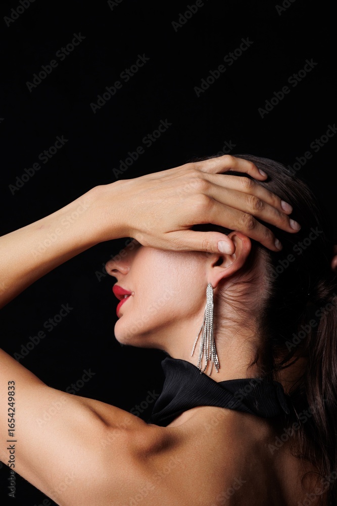 Obraz premium Beautiful young woman in a black elegant dress posing in studio on black background. Fashion model with long hair, red lips, and stylish earrings. Glamour portrait, studio photoshoot, beauty 