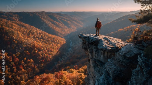 A hiker with a backpack standing at the edge of a cliff overlooking a valley filled with autumn foliage