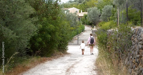 Establishing shot of a woman and her young son walking toward the gate of a stone house surrounded by Mediterranean vegetation in Soller, Mallorca