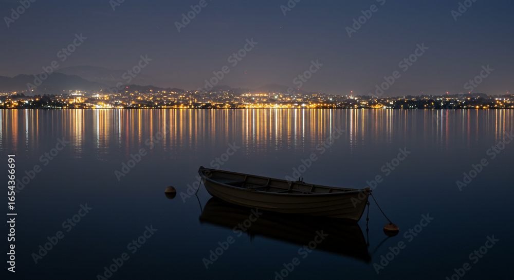 A small boat rests on calm water, reflecting city lights at night under a dark blue sky. A small boat rests on calm water, reflecting city lights at night under a dark blue sky.