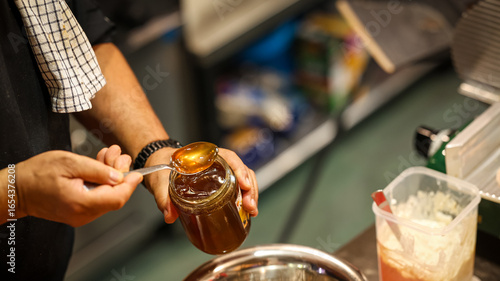 Close-up of person holding a jar of honey with spoon, golden sweet syrup dripping in kitchen scene, concept of natural sweetener and food preparation
