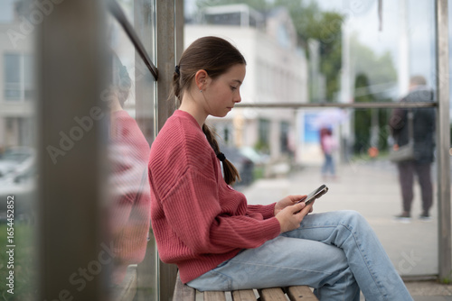 Obraz na plátně Young girl sitting at bus stop, scrolling through social media feed