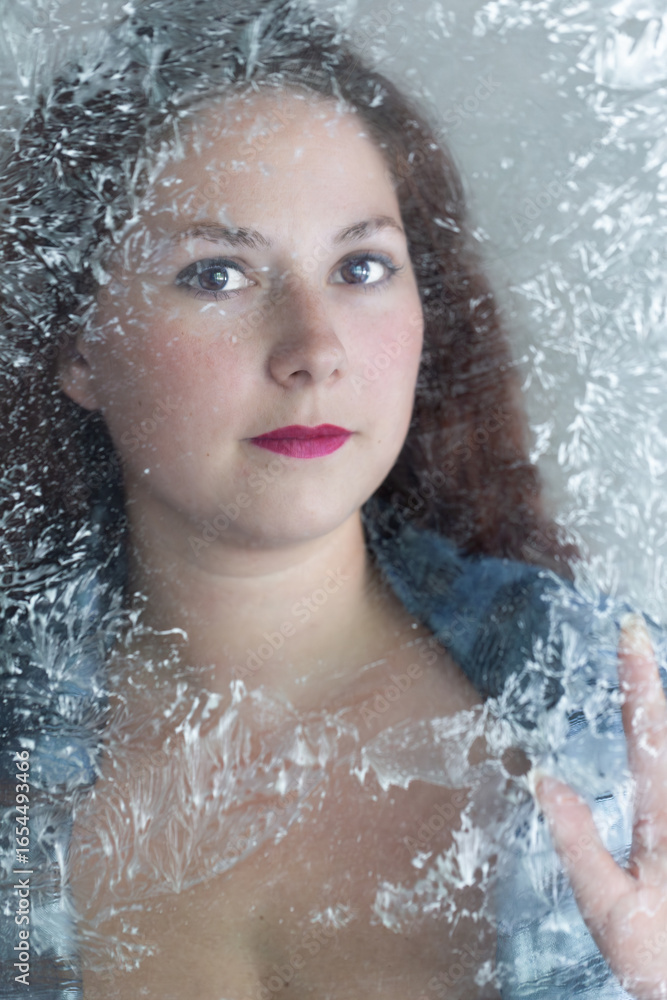 Fototapeta premium Selective focus portrait of beautiful young woman with dark red hair looking through a frosted window 