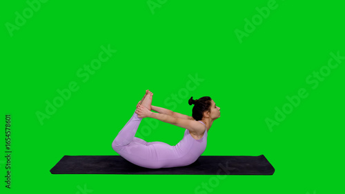 Young sporty fit woman in bodysuit doing yoga bow pose on mat against green chroma key background