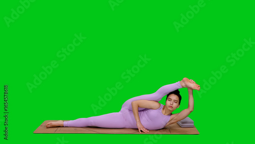 Young sporty woman in bodysuit doing Supta Visvamitrasana pose, Reclining Visvamitra Posture, while practicing yoga on green chroma key background