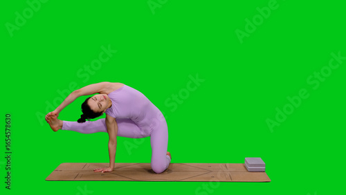 Young sporty woman in bodysuit doing flying warrior pose on mat while practicing yoga on green chroma key background