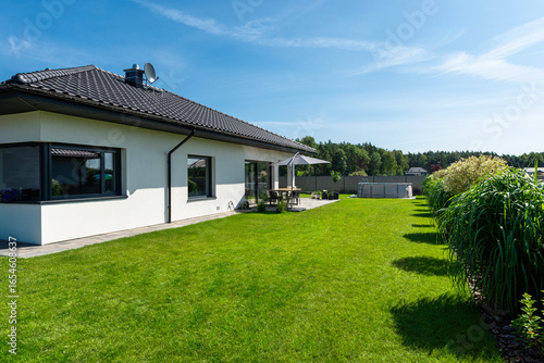 A beautiful garden with green grass, a patio table and umbrella visible, a house in beautiful summer weather, a very wide shot.