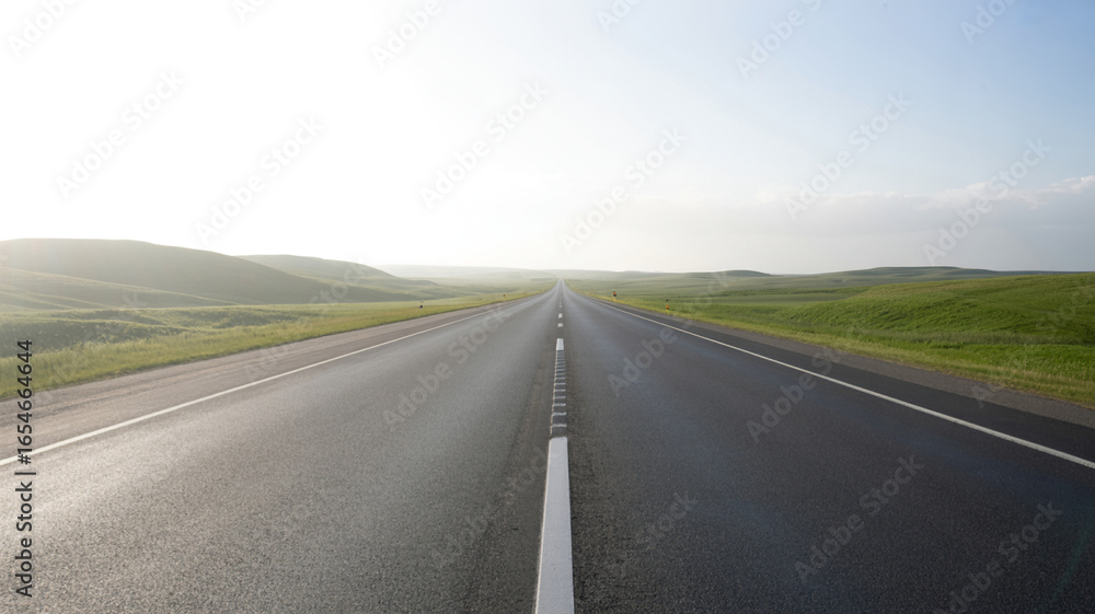 Fototapeta premium An empty asphalt road stretches into a bright hazy horizon with rolling green hills on either side under a pale sky
