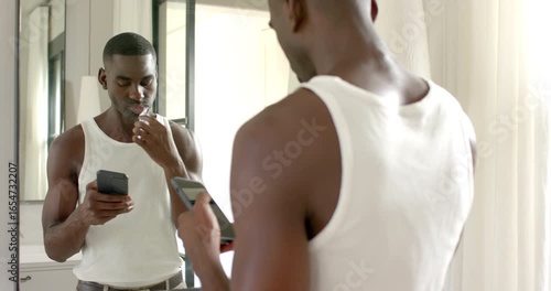 African American man brushing teeth in bathroom, holding smartphone and blue bristle toothbrush