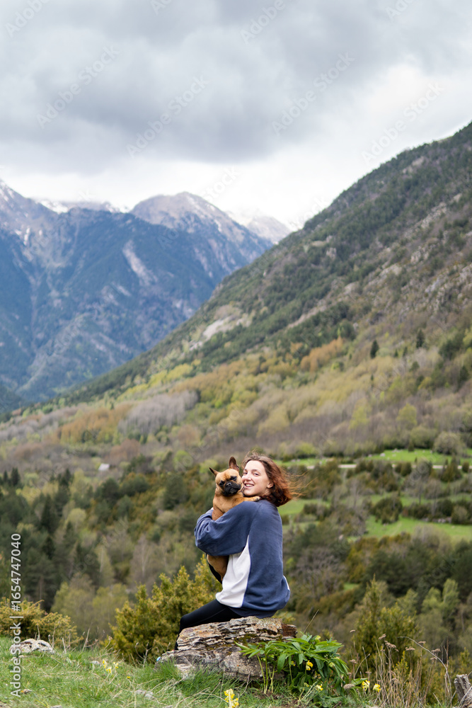 Fototapeta premium Happy traveling woman with cute dog in highlands. Side view of content female hiker sitting on rock and tossing French Bulldog during adventure in Pyrenees mountain range