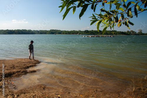 A young boy standing at the edge of a lake, looking out at the water.