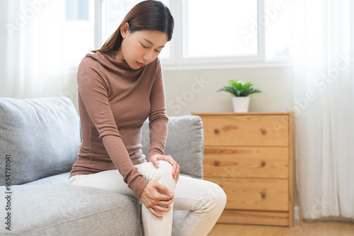 Foto Health care of knee pain concept, suffering asian young woman arthritis sitting joint ache, sore cramp or sprain tendon in leg on sofa at home