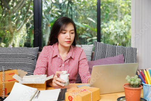 Asian woman working on laptop at home office, holding ceramic product and preparing package for online business shipping, small business owner concept.