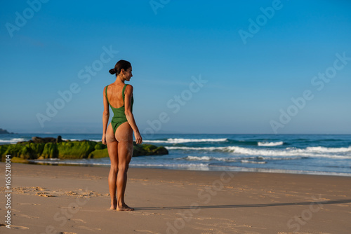 Woman in green swimsuit standing on sandy beach, facing the ocean with hands relaxed, seaweed-covered rocks and waves in background, enjoying peaceful morning light and sea breeze.