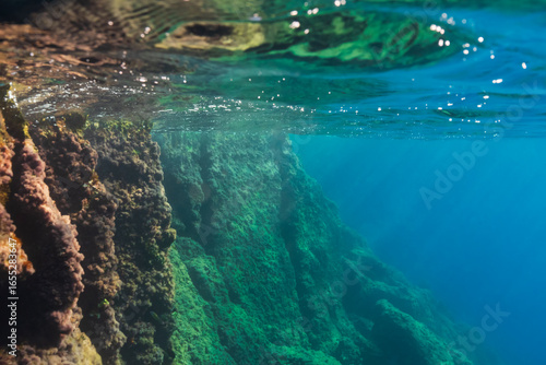 Foto Over-under photograph of a majestic submerged rock wall with sunlight on the surface and a deep blue underwater abyss