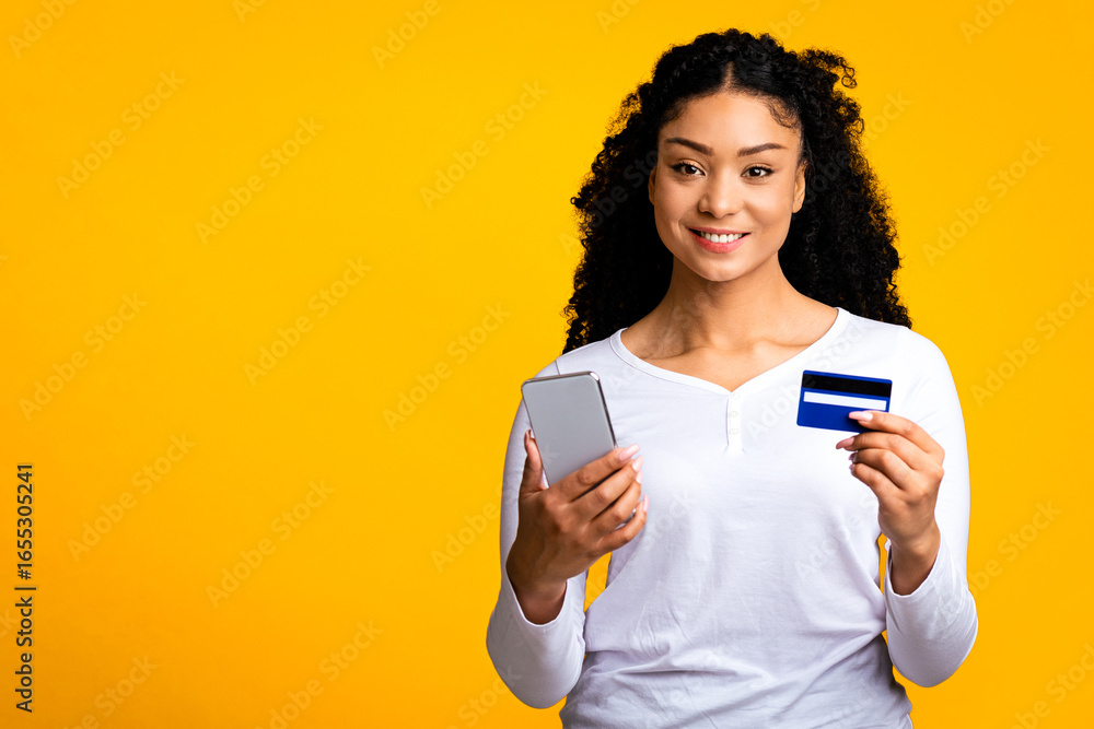 Naklejka premium Portraif of smiling beautiful black woman with smartphone and credit card in hands, young african american female making online shopping, purchasing in internet, standing over yellow background