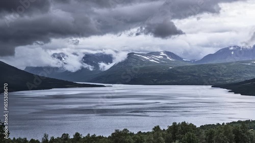 Witness the stunning time-lapse fast-moving dramatic clouds envelop Oppdal lake and surrounding mountains, shifting from vibrant evening hues to a deep night sky. In Trøndelag Norway