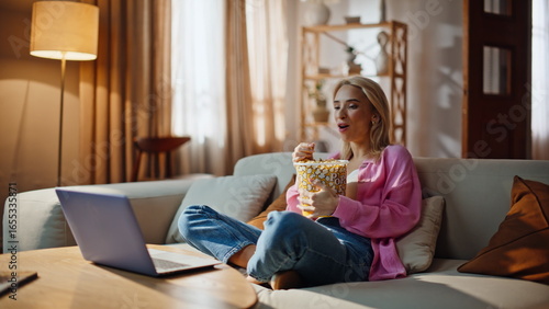 Canvas Print Cheerful blonde streaming series sitcom on laptop snacking chips in comfy house