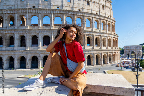 A stylish young woman elegantly sits on a ledge, gracefully posing while showcasing the iconic Colosseum in Rome, framed beautifully against a clear and vibrant blue sky that enhances the scene