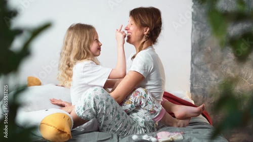 Happy mother and daughter in pajamas sitting on bed, smiling and playing together at home. Cozy family moment, love, care and tenderness, concept of motherhood, childhood and warm relationships.