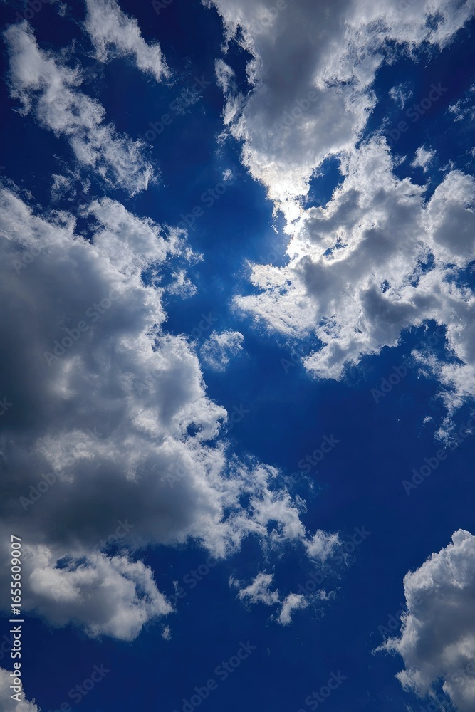 Obraz premium A vibrant, high-angle view of a partly cloudy sky. Bright sunlight peeks through fluffy white and grey clouds against a deep blue backdrop