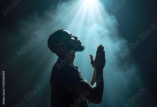 Young black man praying in the dark room with his hands folded in prayer, a beam of light shines on him from above