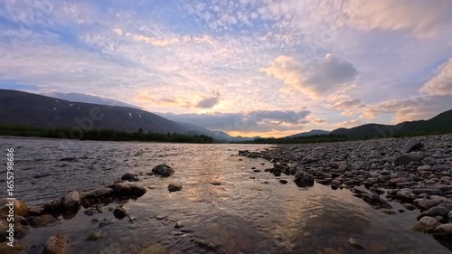 Sunset Time Lapse, a serene river flows over smooth stones with a colorful sky filled with clouds. The mountains create a stunning backdrop to this tranquil scene, inviting reflection and peace.