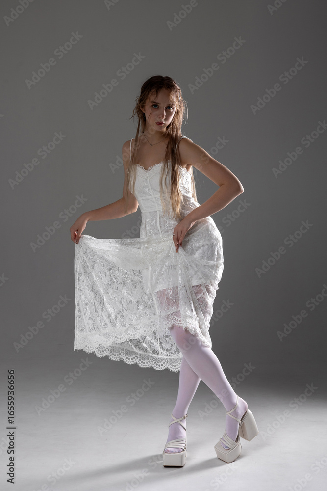 Naklejka premium Young woman poses elegantly in a white lace dress, showcasing a unique style with graceful expression and modern footwear in studio