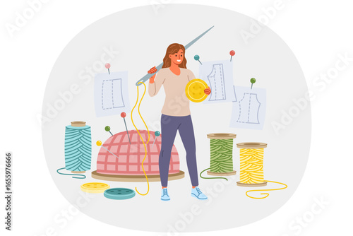 Woman seamstress holds large needle and button, standing near threads and cutting sheets for making handmade clothes. Girl seamstress smiles enjoying creative work of developing dress designs