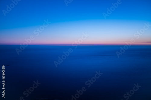 Minimal aerial seascape at dawn with soft pink and blue gradient over calm ocean horizon, showing peaceful twilight colors and infinite natural tranquility