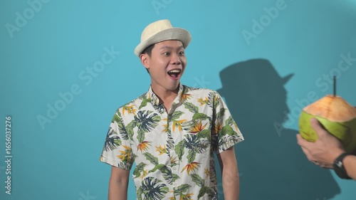 An Asian person drinking from a coconut and making an inviting gesture, filmed against a simple background, symbolizing tropical refreshment, hospitality, lifestyle, and vacation vibes