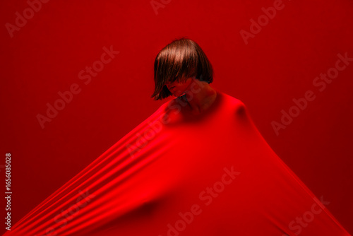 A woman with short hair is covered by a taut red fabric, creating strong lines and shadows. Her head is tilted down against the vibrant red background. This abstract composition highlights form.