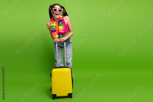 Young stylish woman ready for vacation with bright yellow suitcase and vibrant lei necklace on green background