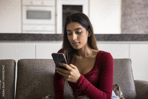 Digital connection. Serious young zoomer woman sit on couch at home focused on mobile phone screen text important message read news online wait for call check business email engaged in crypto trading
