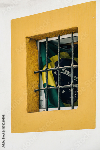 A beautiful Brazilian flag is positioned behind the bars of a window, set against a brightly painted yellow wall. The scene reflects local pride and cultural identity.