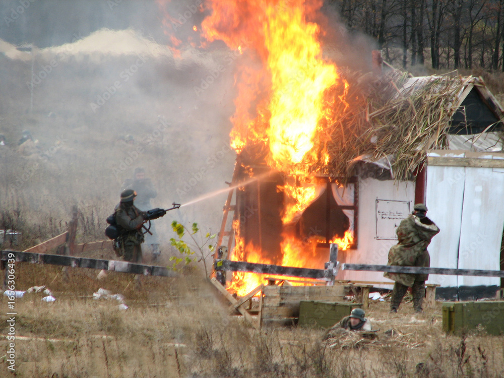 Fototapeta premium Historical reenactment of World War II battle with soldier using flamethrower to set wooden hut on fire, symbolizing wartime combat, destruction, military history and battlefield atmosphere