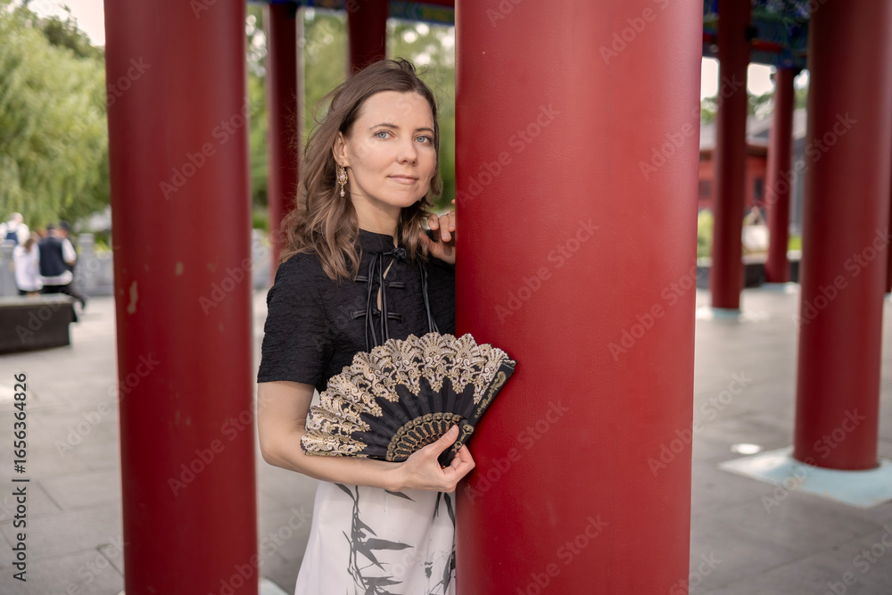 Obraz premium Portrait of a stylish woman in a black and white dress holding an ornate lace fan while leaning against a large red column in a traditional oriental pavilion