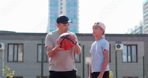 Father teaching son basketball on outdoor court. Parent coaches child in slow motion. Dad shows boy shooting skills. Family bonding, sports practice, and mentorship create joyful learning