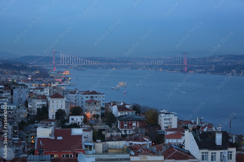 Obraz premium The illuminated Bosphorus Bridge connecting Europe and Asia at twilight, with boats crossing the strait and Istanbul’s city lights glowing.