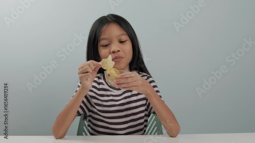 A cute kid girl in a patterned shirt is eating seaweed potato chips from a small cup. She is happy to have her favourite snack during her holiday. plain background