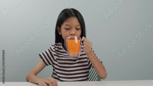 A cute girl wearing a patterned shirt is sitting and drinking a glass of orange juice with delight. She smiles and gives a thumbs up in appreciation. plain background