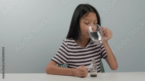 A cute girl in a patterned shirt and long black hair lifts a glass of water on a white table and takes a sip after eating a small bottle of medicine nearby. The background is plain.