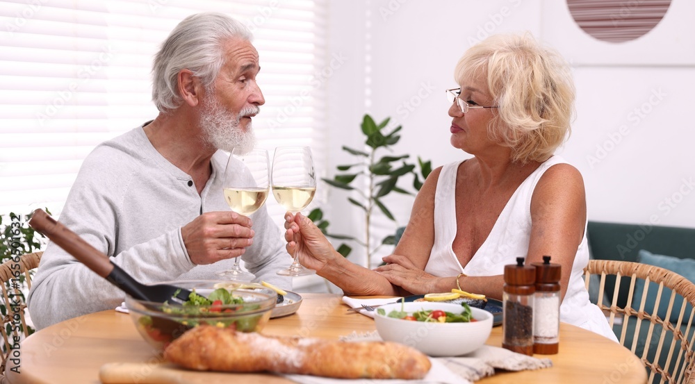 Elderly couple clinking glasses of white wine during dinner at table indoors Elderly couple clinking glasses of white wine during dinner at table indoors