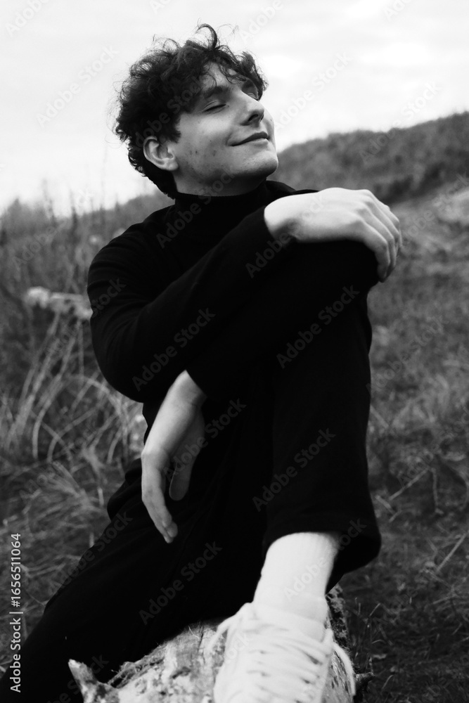 Fototapeta premium Emotion black and white portrait of a young guy in a black wear. Man have relax and smile. Seating on the tree